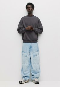 Šedá mikina s rebrovanými manžetami, spojená so svetlomodrými voľnými cargo džínsami s viacerými vreckami a jemnou textúrou denimu. Outfit dopĺňajú tenisky.