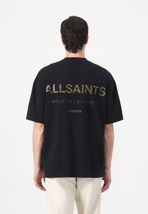 Man draagt een zwarte oversized t-shirt met "ALLSAINTS" en Japanse tekens in camouflageprint en "LONDON" in goud op de rug.
