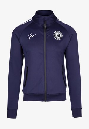 Marineblauwe sportjack met hoge kraag, volledige ritssluiting, zijzakken en witte accenten; voorzien van een cirkelvormig logo op de borst.