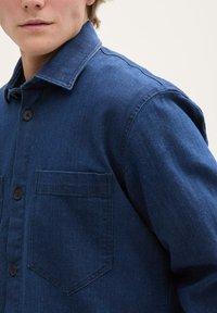 Jeansowa koszula w ciemnym niebieskim kolorze, z ostrym kołnierzykiem, z przodem zapinanym na guziki oraz dwiema kieszeniami na piersi. Tkanina ma teksturowane wykończenie z subtelnymi punkcikami.
