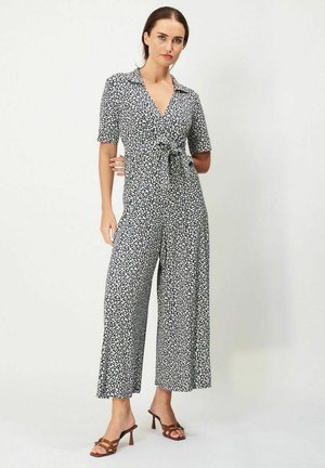 Sort-hvid blomstret jumpsuit med krave, korte ærmer, frontsnøring og vidde ben, parret med brune hæle sandaler.