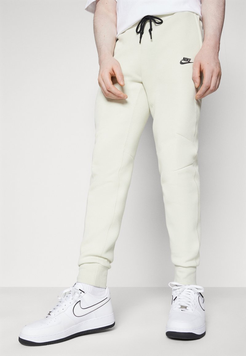Helle cremefarbene Nike-Sweatpants mit schwarzem Kordelzug und Logo, mit schmal zulaufenden Beinen. Tragen Sie dazu weiße Nike-Sneaker mit schwarzen Akzenten.