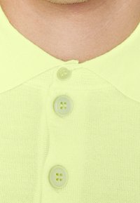 Světle žlutá pletená polo košile se třemi kulatými knoflíky se čtyřmi dírkami na límci. Hladký texture a jednoduchá struktura.