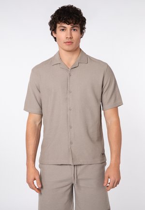 Young man with curly hair wearing a beige short-sleeve button-up shirt and matching shorts against a plain background.