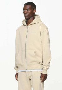 Beige zip-up hette med snor i hetta, frontlommer og subtil logo detalj. Laget av myk stoff med en avslappet passform.