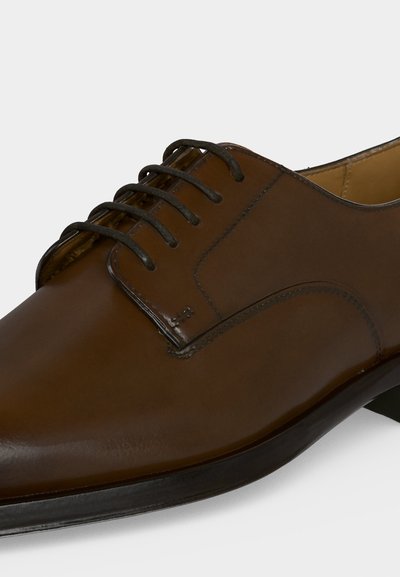 Chaussure Oxford formelle en cuir marron avec des lacets noirs et un bout légèrement pointu sur fond blanc.