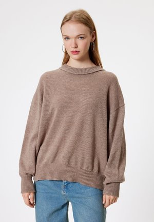 Beżowy sweter w oversize z prążkowanym kołnierzem i mankietami, o miękkiej fakturze. W połączeniu z jasnoniebieskimi dżinsami z wysokim stanem.