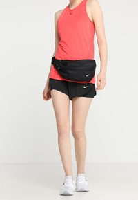 Musta vyölaukku, jossa on kaksi vetoketjullista osastoa, päällä kirkkaan pinkin hihattoman paidan ja mustien shortsejen kanssa. Valkoiset lenkkitossut täydentävät asun.