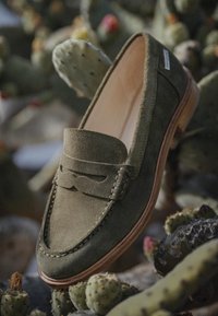Mocassini in suede verde con design cucito, punta stile mocassino e suola in gomma beige, posizionati su uno sfondo di cactus.