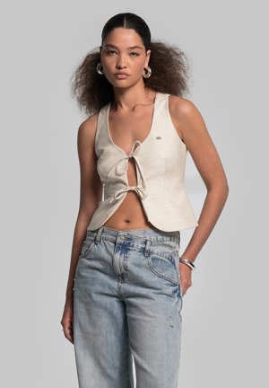 Top corto beige con design a cravatta, senza maniche e con un piccolo logo. Abbinato a jeans in denim blu chiaro, a vestibilità ampia.