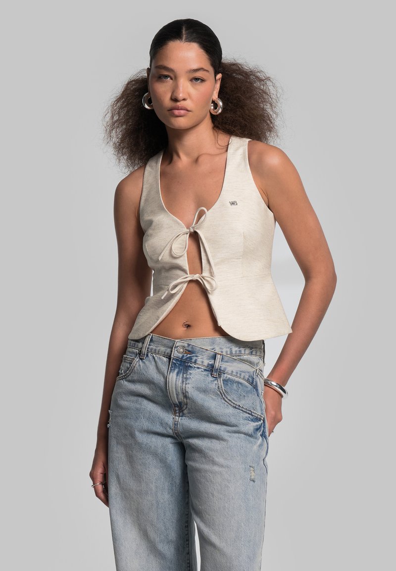 Beiger Crop-Top mit einer Krawatten-Frontgestaltung, ärmellos und mit kleinem Logo. Kombiniert mit hellblauen, locker sitzenden Denim-Jeans.