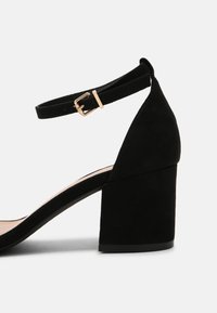 Svart mocka klackskor med blockklack och ankelrem, pryds av en guldspänne. Slät yta och öppna sidor för andningsförmåga.