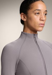 Svetlosivý športový kabát s vysokým golierom, plným predným zipsom a textúrovanou látkou, ktorá obsahuje šitie na pleciach.