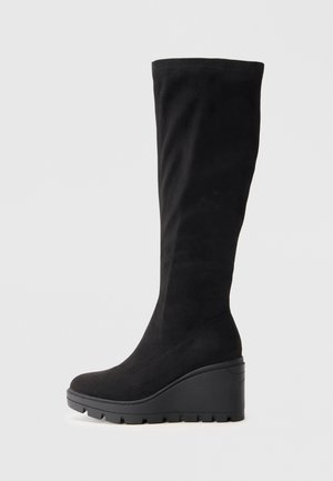 Black suede knee-high boot with a wedge sole, featuring a rounded toe and textured finish; smooth design without embellishments.