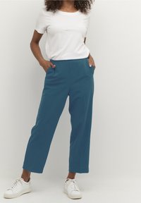 Teal, losvallende broek met zijzakken, gemaakt van een gladde stof; gecombineerd met een eenvoudige witte t-shirt met ronde hals en witte sneakers.