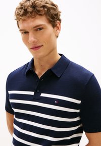 Marineblå polo med hvide vandrette striber, korte ærmer og krave. Lille broderet logo på brystet. Glat tekstur.