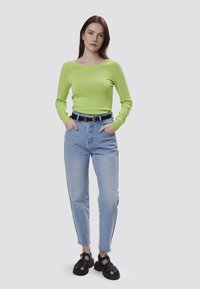 Maglia verde lime a maniche lunghe, jeans azzurri a vita alta e sandali neri con suola spessa. La modella posa con le mani in tasca.
