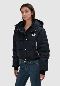 Czarna krótka kurtka puchowa z kapturem, zapinana na zatrzaski, z wyraźnym logo na piersi. Napis na boku.