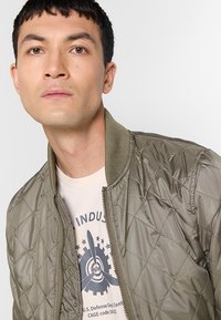 Gestepptes olivgrünes Bomberjacke mit geripptem Kragen und Reißverschluss vorne. Getragen über einem hellfarbenen T-Shirt mit mechanischem Grafikdesign.