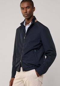 Chaqueta ligera de color marino con cuello alzado, cierre de cremallera, dobladillo elástico y bolsillos laterales. Presenta un acento beige en el cuello.