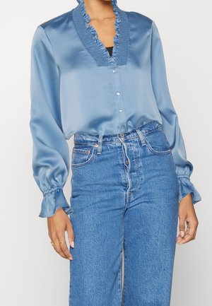 Kobieta w jasnoniebieskiej satynowej bluzce z marszczonym kołnierzem i mankietami, w połączeniu z niebieskimi dżinsami o średnim odcieniu i wysokim stanie.