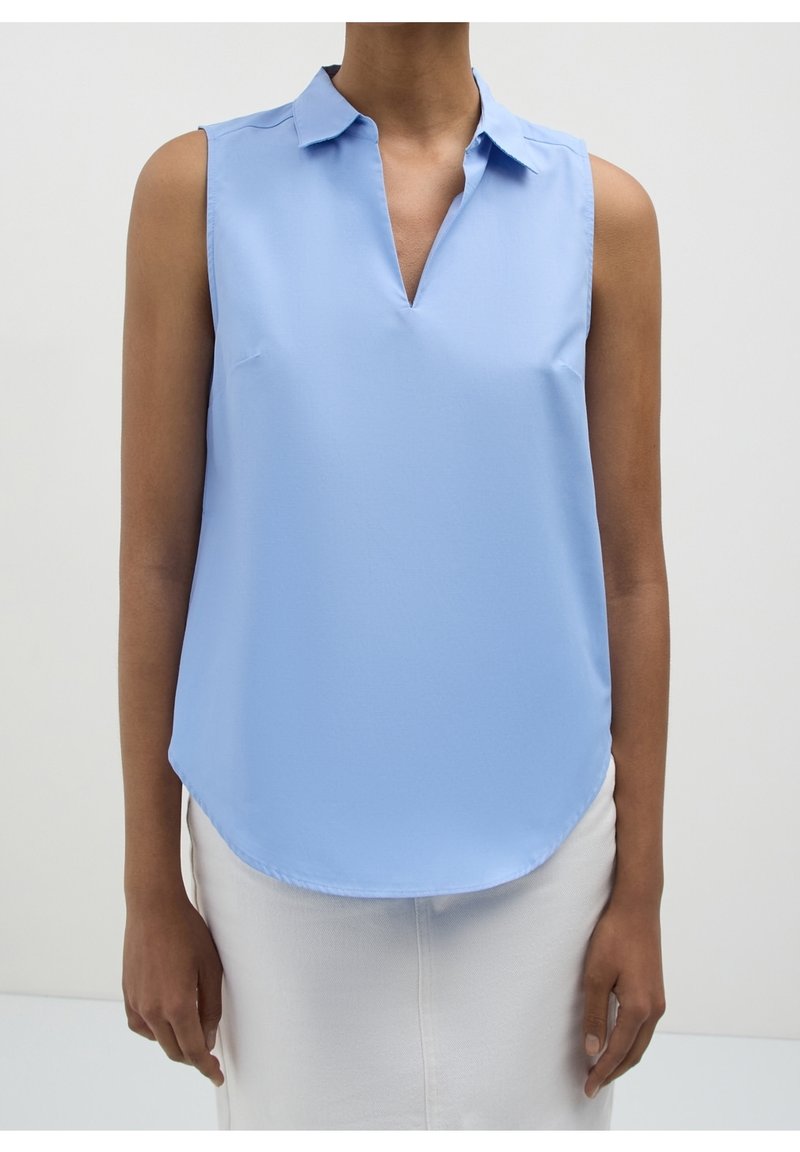 Blouse sans manches bleu clair avec col et encolure en V portée par une personne portant un pantalon clair sur un fond uni.