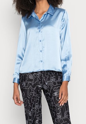 Femme portant une chemise en satin bleu clair boutonnée et un pantalon noir avec des motifs abstraits de lignes blanches.