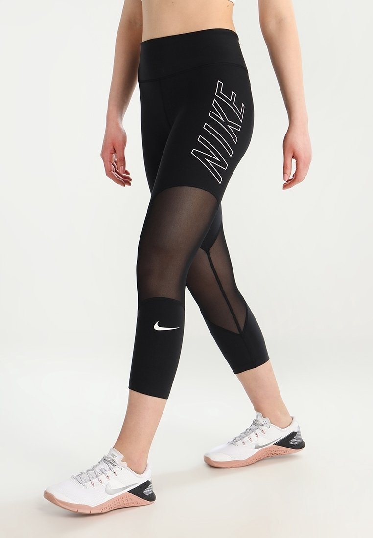 Černé legíny s vysokým pasem, s mesh panely a bílým logem "NIKE" na boku. Nošeny s bílými a růžovými sportovními botami.