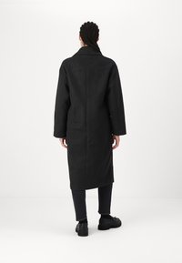 Cappotto di lana nera con un design di lunghezza media, colletto largo e silhouette dritta, abbinato a pantaloni scuri e scarpe nere.