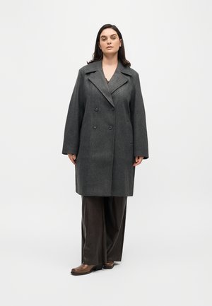 Femme debout face à l'objectif portant un manteau trench-coat gris foncé à double boutonnage, un pantalon large foncé et des chaussures marron sur un fond uni.