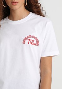 Bílý bavlněný tričko s zakřiveným červeným textem: "DREAM DÖNER TASTY & FRESH" na levé straně. Krátké rukávy, klasický střih s kulatým výstřihem.