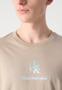 Bežinė medvilninė marškinė su apvaliu kaklu, su balta ir akvamarinine logotipo išvaizda, esanti krūtinės srityje. Lygus paviršius.