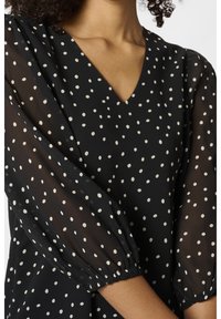 Blouse noire à pois blancs avec col en V et manches bouffantes semi-transparentes. Présente des pois blancs et des poignets élastiques pour un ajustement confortable.