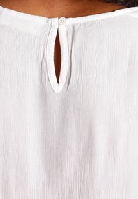 Blusa bianca con texture, caratterizzata da un collo rotondo con un'apertura a forma di chiave e una chiusura con un solo bottone sul retro.