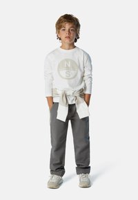 Camicia bianca a maniche lunghe con un logo circolare e pantaloni grigi. La camicia ha un felpa beige annodata in vita, completata da sneakers grigie.