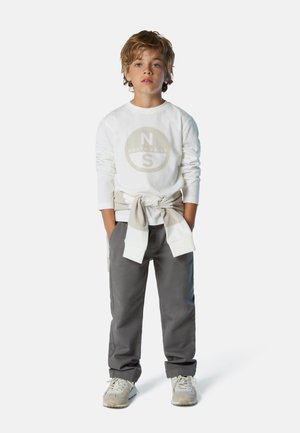 Camicia bianca a maniche lunghe con un logo circolare e pantaloni grigi. La camicia ha un felpa beige annodata in vita, completata da sneakers grigie.