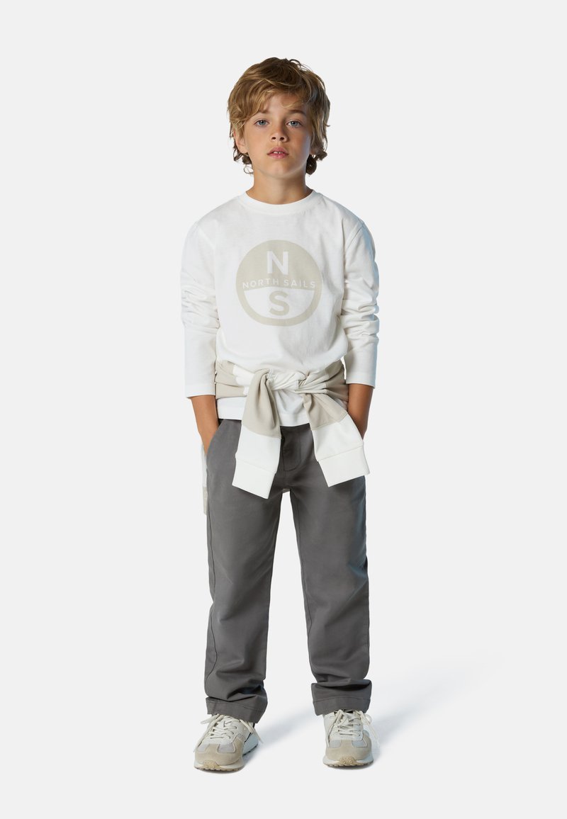 Camicia bianca a maniche lunghe con un logo circolare e pantaloni grigi. La camicia ha un felpa beige annodata in vita, completata da sneakers grigie.