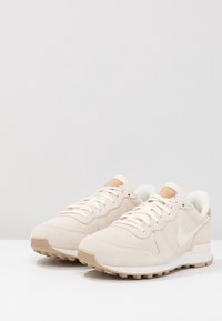 Beiga mocka sneakers med en vit läderswoosh, texturerad yta och ljusbruna accenter på hälen. Gummisula med mönster för bra grepp.