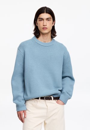 Jeune homme aux cheveux foncés mi-longs porte un pull en maille bleu clair et un pantalon crème, debout avec une main dans la poche sur fond blanc.