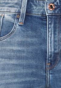 Jeans de mezclilla en azul claro con una textura desgastada. Presentan un botón de cobre, un solo bolsillo frontal y costuras en naranja contrastante a lo largo de las costuras.