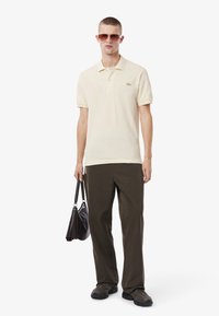 Beige pikétröja med korta ärmar och en grön logotyp. Mörkgröna byxor och svarta sneakers. Brun handväska med axelrem.