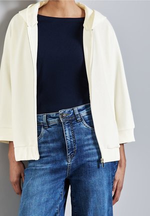 Persona que lleva una sudadera blanca con cremallera sobre una camiseta negra y vaqueros azules de talle alto, sobre un fondo gris liso.
