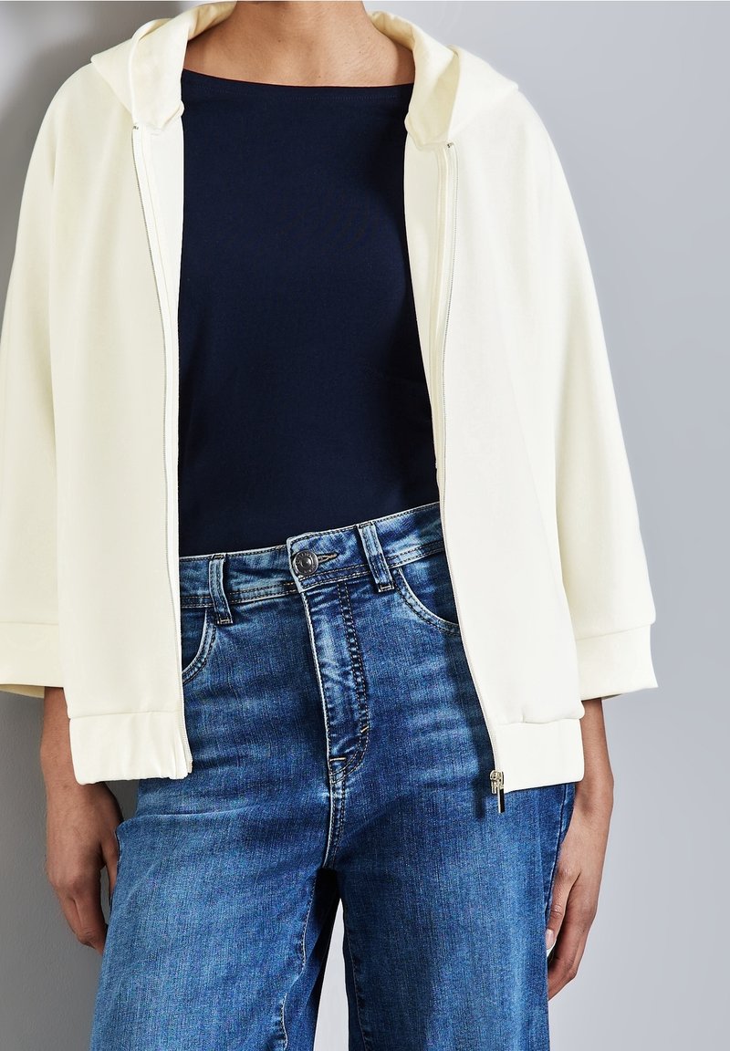 Personne portant un sweat à capuche blanc zippé par-dessus un top noir et un jean bleu taille haute sur un fond gris uni.