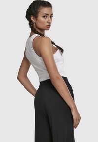 Femme aux cheveux tressés portant un haut blanc sans manches et un pantalon noir, regardant par-dessus son épaule sur un fond uni.