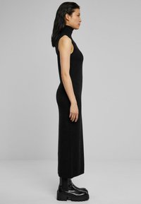 Robe noire sans manches avec col roulé, au silhouette ajustée, fabriquée dans un matériau lisse et extensible. Assortie à des bottes noires imposantes.