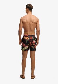 Homme debout pieds nus portant un short de bain à motif floral et des sandales marron, vu de dos sur un fond blanc.