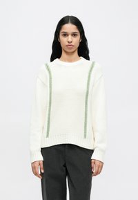 Bijeli pleteni džemper sadrži zelene šavove uz bočne strane te rebrasti ovratnik i rub. Uparen sa sivim suženim hlačama.