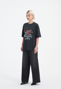 Schwarzes, übergroßes T-Shirt mit einem Grafikdesign aus Totenkopf und Herz, kombiniert mit weit geschnittenen dunklen Jeans und spitzen schwarzen Schuhen mit Kettenakzenten.
