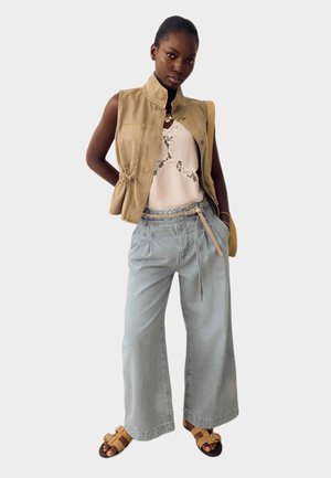 Kvinde iført ærmeløs beige vest, lyserød blonde top, vide lyseblå jeans med bælte og brune sandaler, stående foran hvid baggrund.