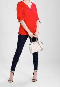 Punainen lyhythihainen paita, jossa on V-pääntie ja solmukohtia, yhdistettynä tummiin tiukkoihin farkkuihin ja mustiin remmikenkiin. Pieneen vaaleanpunaiseen käsilaukkuun on tartuttu.